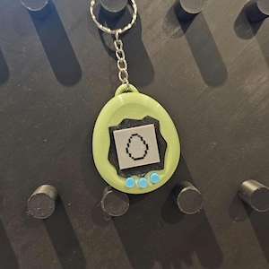 May include: A green and black keychain resembling a retro digital pet game. It features a silver chain and ring. The screen displays a pixelated egg design, with three blue buttons beneath.