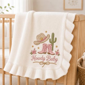 May include: A cream-colored knit baby blanket with a ruffled edge featuring an embroidered design of a tan cowboy hat, pink cowboy boots, a green cactus, small gold stars, and pink flowers. The text Howdy Baby is embroidered in pink script.