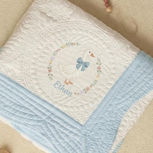 Op de afbeelding: Witte en lichtblauwe babyquilt met een ganzenontwerp. De quilt heeft een witte gans met een blauwe strik, omringd door een bloemenkrans. De naam "Ethan" is lichtblauw geborduurd. De quilt heeft een gewatteerd patroon en een lichtblauwe rand.