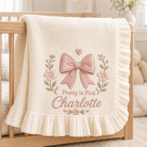 Könnte beinhalten: Eine cremefarbene Strickdecke mit gerüschtem Rand und gestickten Details. In der Mitte befinden sich eine rosa Schleife, ein kleines Herz und Blumenzweige. Der Text lautet Pretty in Pink und der Name Charlotte in rosa Schreibschrift.