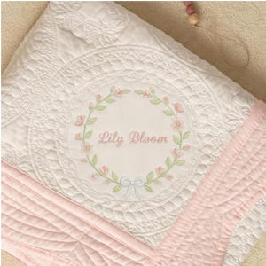 Puede incluir: Manta de bebé acolchada blanca con borde rosa y diseño de corona floral. El centro presenta el texto "Lily Bloom" en escritura rosa. La manta tiene un patrón detallado y texturizado.
