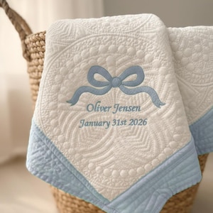 Puede incluir: Una manta de bebé acolchada blanca con un lazo y ribete azul claro. La manta presenta el nombre bordado "Oliver Jensen" y la fecha "31 de enero de 2026". La manta está doblada y colocada en una cesta de mimbre.