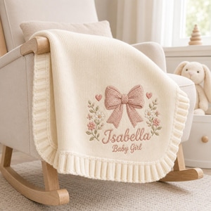 May include: A cream-colored knitted baby blanket featuring a ribbed border and central embroidery. The design includes a large dusty pink bow, small floral sprigs, two hearts, and the embroidered text Isabella Baby Girl in a script font.
