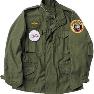 May include: Olive green military-style jacket with a zipper closure and multiple pockets. Features a small patch with a gold emblem and a round button with the text "We Are The People." A "King Kong Company" patch is on the right sleeve.