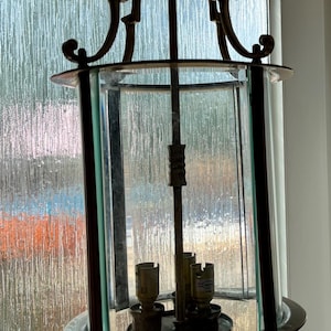 May include: An antique brass chandelier with a cylindrical glass enclosure. The fixture has a decorative top and bottom, with three light bulb sockets visible inside. The glass panels are framed by dark metal.