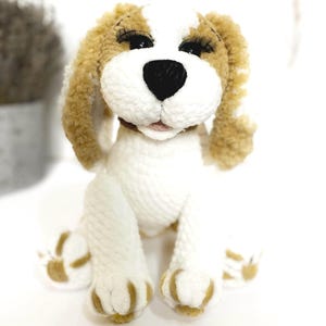 May include: A handcrafted, plush dog toy in a seated pose. The toy has a white body with tan ears, paws, and details. It features a black nose, long eyelashes, and a small brown collar. The dog is made of soft, textured yarn.