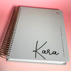 May include: A light gray spiral-bound book with gold-colored coils. The cover features the name "Kara" in black script and the text "THE NEW TESTAMENT | CHRISTIAN STANDARD BIBLE". A small gold icon is in the upper right corner.