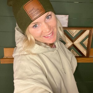 Puede incluir: Un gorro verde oliva con un parche de cuero marr&oacute;n que dice "MAKE. HEAVEN. CROWDED." El gorro se usa con una sudadera con capucha color crema. El fondo presenta una pared verde y una obra de arte de madera.