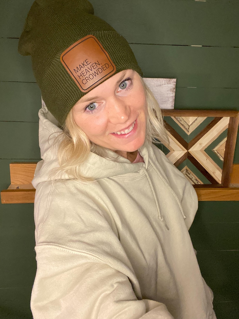 Puede incluir: Un gorro verde oliva con un parche de cuero marr&oacute;n que dice "MAKE. HEAVEN. CROWDED." El gorro se usa con una sudadera con capucha color crema. El fondo presenta una pared verde y una obra de arte de madera.