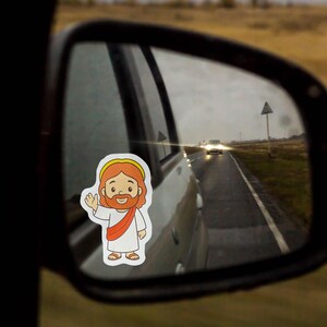 May include: A sticker of a cartoon Jesus waving, with red hair, a beard, and a white robe, is affixed to a car's side mirror. The mirror reflects a road with a car driving on it under a cloudy sky.