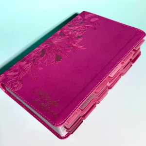 May include: A fuchsia-colored Bible with a debossed floral pattern on the front cover. The cover features gold-colored cursive text and includes integrated side tabs labeled with book names such as Genesis, Exodus, Leviticus, Numbers, and Samuel.