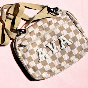 May include: A beige and white checkered crossbody bag featuring a zippered front pocket and an adjustable tan strap with black plastic hardware. The name AVA is displayed on the front in large, white, textured, varsity-style letter patches.