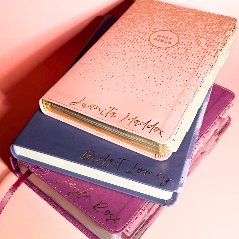 May include: A stack of three personalized Holy Bibles with gold foil lettering. The top book is light pink with a gold dot pattern, the middle is navy blue, and the bottom is magenta. Each cover features a custom name in elegant cursive script.