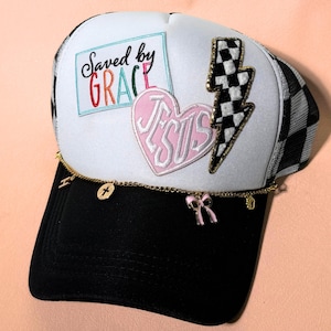 May include: A black and white trucker hat with a white front panel featuring the text "Saved by GRACE" and a pink heart with "JESUS" embroidered on it. A gold chain with charms hangs from the brim. A checkered lightning bolt is on the side.