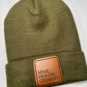 Puede incluir: Gorro verde oliva con un parche de cuero marr&oacute;n. El parche presenta el texto "MAKE. HEAVEN. CROWDED." en negrita. El gorro est&aacute; hecho de un material de punto suave y tiene un borde doblado.