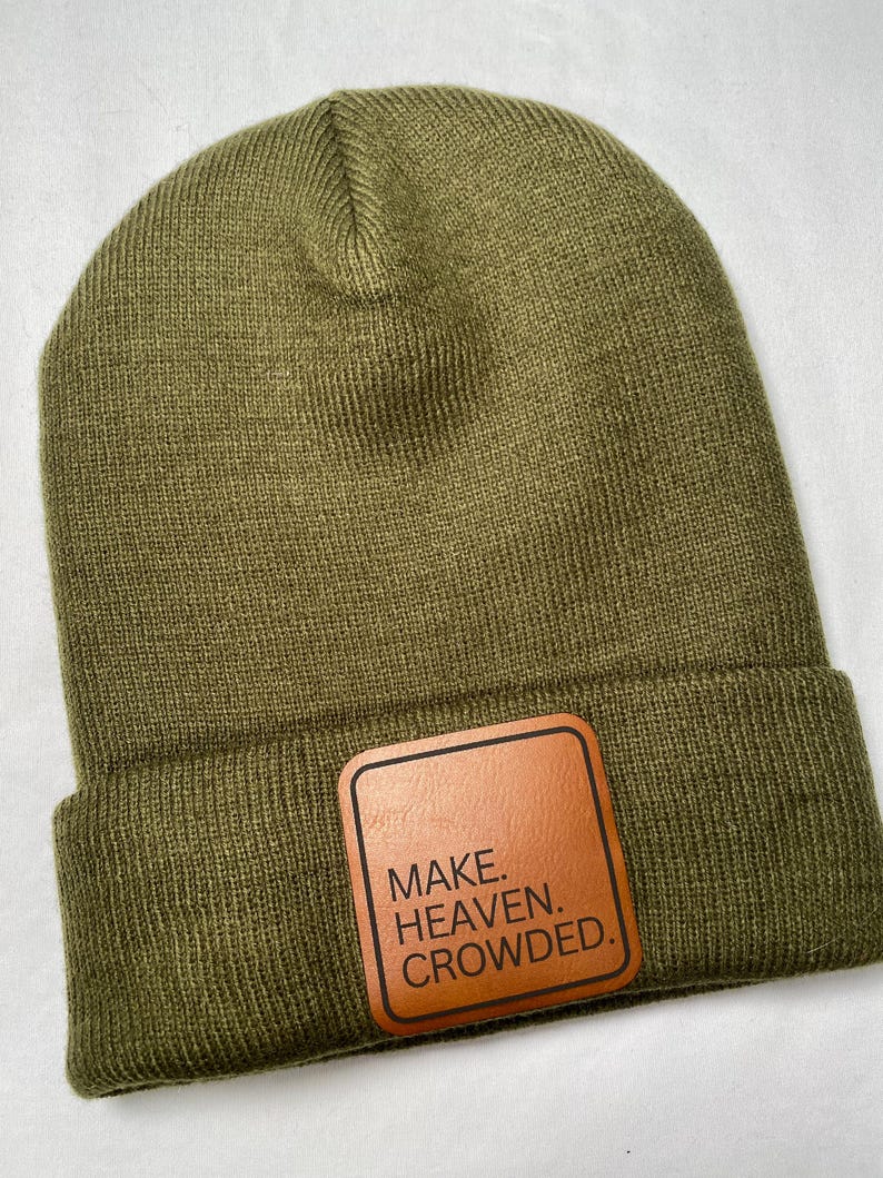 Puede incluir: Gorro verde oliva con un parche de cuero marr&oacute;n. El parche presenta el texto "MAKE. HEAVEN. CROWDED." en negrita. El gorro est&aacute; hecho de un material de punto suave y tiene un borde doblado.
