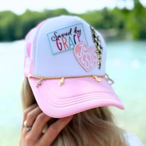 May include: A pink and white trucker hat with the text "Saved by GRACE" and a heart-shaped "JESUS" patch. The hat features a gold chain with charms and a pink brim. The hat has a mesh back and a curved brim.