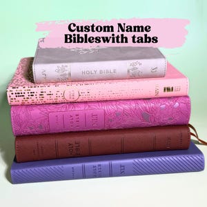 May include: A stack of five Bibles with varying covers: a light gray one with floral details, a pink one with gold dots, a magenta one with floral embossing, a dark brown one, and a purple one with diagonal lines. Text reads: Custom Name Bibleswith tabs.