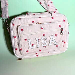 May include: Cream-colored corduroy shoulder bag with a repeating pattern of red strawberries. The bag features a front pocket with the name "LISA" in silver glitter letters. Includes a matching strap and gold-toned hardware.