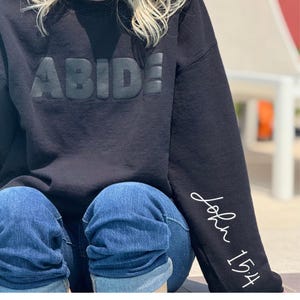May include: Black sweatshirt with the word "ABIDE" in raised lettering across the chest. The sleeve features the text "John 15:4" in a handwritten style. The person is wearing blue jeans.