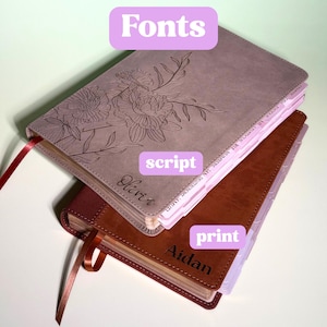 May include: Two rectangular journals with leather-like covers. The top journal is mauve with an engraved floral design and the name Olivia in script. The bottom journal is brown with the name Aidan in print. Both have ribbon bookmarks and tabs.