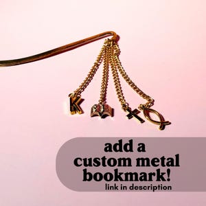 May include: A gold-colored metal bookmark featuring four dangling chains with charms: the letter K, an open book, a cross, and a fish symbol. The text "add a custom metal bookmark! link in description" is overlaid at the bottom of the image.