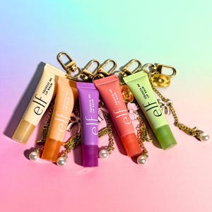 May include: An assortment of colorful lip balm tubes with gold-colored keychains and charms. Each tube is a different color, including yellow, orange, purple, red, and green. The text "Squeeze Me Lip Balm" and the brand "elf" are printed on each tube.