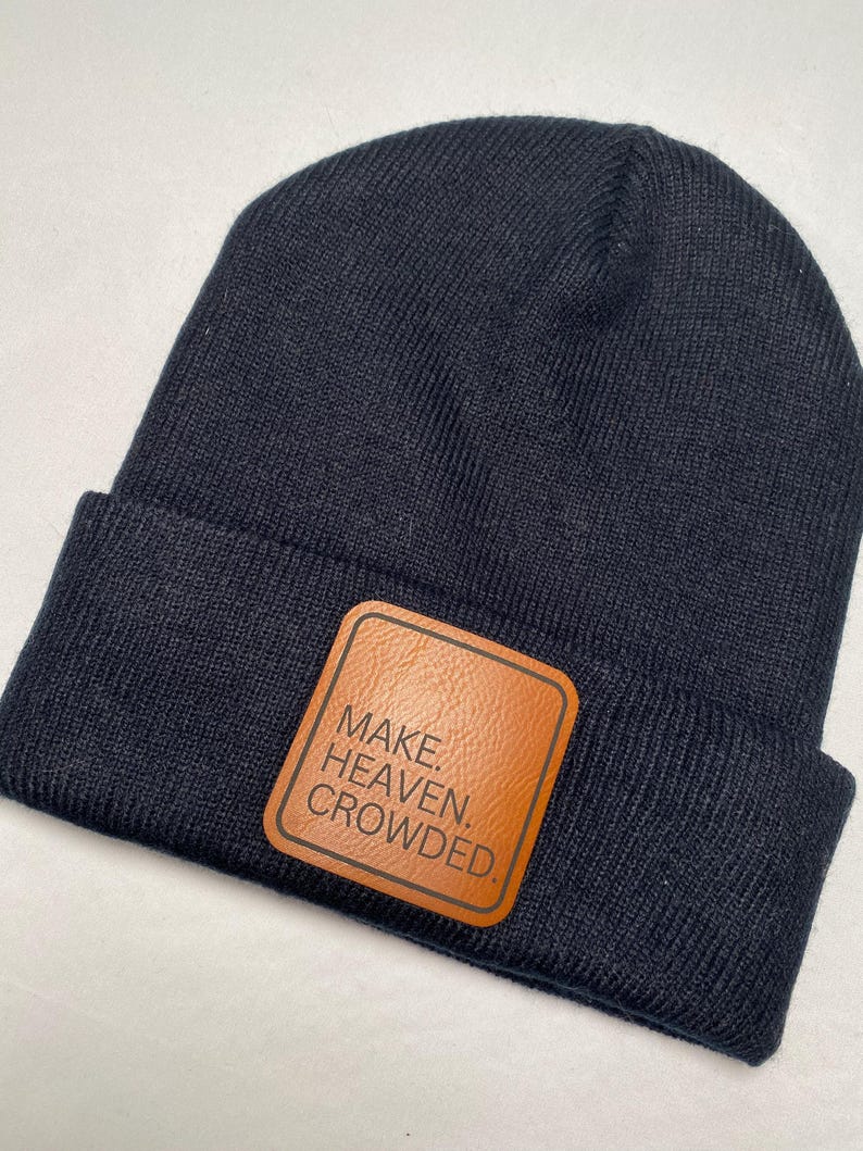 Puede incluir: Un gorro de punto azul marino con un parche de cuero marr&oacute;n. El parche presenta las palabras "MAKE. HEAVEN. CROWDED." en letras blancas. Un estilo de gorro cl&aacute;sico, adecuado para el clima fr&iacute;o.