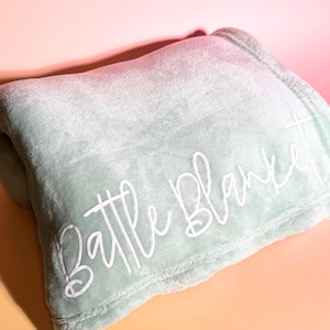 May include: A soft, mint green blanket with the words "Battle Blanket" in white script. The blanket is folded and appears plush, with a cozy texture. The background has a gradient of pink and orange.