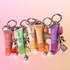 May include: Six tubes of lip balm in various colors, each attached to a keychain. The tubes are labeled "Squeeze Me Lip Balm" by "elf". Each keychain has a charm, including gummy bears, pearls, and small decorative bottles.