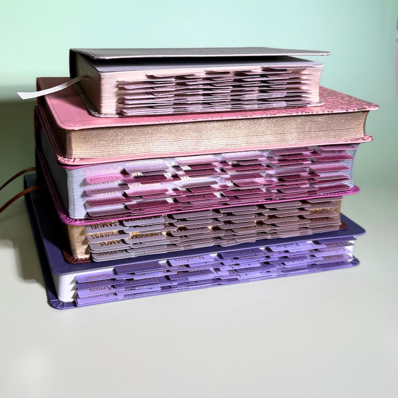 May include: A stack of four books with indexed tabs along the edges. The top book is light gray, followed by a pink book with gold-gilded edges, a book with pink and purple tabs, and a bottom book with purple tabs and a purple cover.
