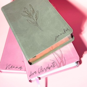 May include: A sage green journal with a leaf illustration and the name Jennifer engraved on the cover, stacked atop a pink journal featuring the names Vienna and Ava Elizabeth with a linear plant design, both with visible page edges.