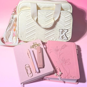 May include: Cream-colored quilted handbag with a shoulder strap and a large letter "K" patch. Pink notebooks, pens, and a highlighter are arranged with gold paper clips. A tassel with beads and charms hangs from the bag.