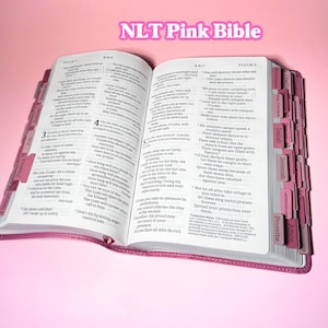 May include: An open NLT Pink Bible with white pages featuring black printed text. The book has pink index tabs along the edges labeled with book names like Psalms, Isaiah, and Proverbs. The cover is a textured pink material.