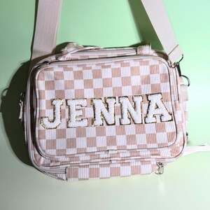 May include: A rectangular crossbody bag featuring a light pink and white checkered pattern. The front displays the name JENNA in large, white, textured varsity-style letters with gold glitter outlines. The bag includes a matching adjustable strap.