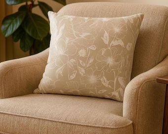 May include: A beige square throw pillow with a floral pattern in white, resting on a beige armchair. The pillow features a detailed design of flowers and leaves, adding a touch of elegance to the room's decor.