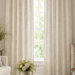 May include: A pair of floor-length curtains in a light beige color featuring a subtle, tone-on-tone damask pattern. The fabric hangs in soft, vertical folds, covering the sides of a window that allows natural light to filter through the center.
