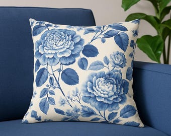 May include: Square throw pillow with a cream background and a blue floral pattern. The design features large, detailed blue roses and leaves, creating a classic, elegant look. The pillow is placed on a blue sofa.