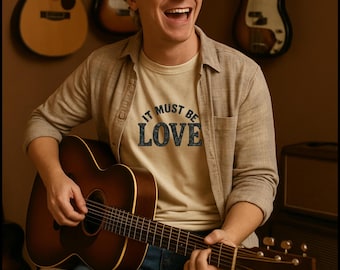 May include: A man wearing a beige t-shirt with the text "It Must Be Love" printed on it, is sitting on a stool and playing an acoustic guitar. He is smiling and looking at the camera. There are two guitars in the background.