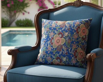 Puede incluir: Un cojín azul con estampado floral descansa sobre un sillón azul tapizado con detalles de madera oscura. El cojín presenta un diseño vibrante con flores rosas, amarillas y burdeos sobre un fondo azul. La silla está cerca de una ventana con vista a una piscina.