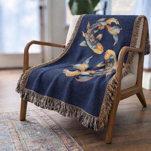 May include: A navy blue throw blanket draped over a chair, featuring a koi fish design in shades of orange, yellow, and blue. The blanket has a fringed edge in a complementary color, adding texture and visual interest. The chair has a wooden frame.