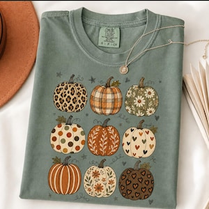 Peut inclure: Un t-shirt à manches courtes vert sauge avec une impression graphique de neuf citrouilles disposées en grille trois par trois. Les citrouilles présentent divers motifs dont léopard, carreaux, fleurs, pois, feuilles et cœurs. Col rond.