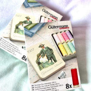 May include: Two boxes of Gütermann Creativ sewing thread. Each box contains eight spools of thread in various pastel colours. The boxes are decorated with a vintage illustration of a couple in traditional clothing. The text on the boxes reads "Gütermann Creativ" and "Nostalgie Box Allesnäher 100 m".
