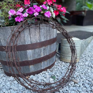 May include: A circular coil of rusty barbed wire rests on a bed of gray gravel. In the background, a weathered wooden barrel planter displays vibrant pink and purple flowers. A metal watering can is also visible.
