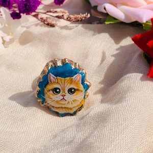 May include: A gold ring with a blue background and a painted image of a ginger cat with white fur. The cat has large, round eyes and a sweet expression.