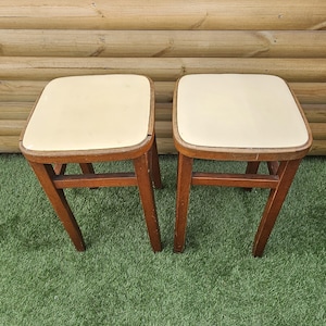 May include: Two square stools with wooden frames and off-white padded seats. Each stool features four straight wooden legs connected by horizontal support bars. The wooden frames show a medium brown finish with visible grain and minor surface wear.