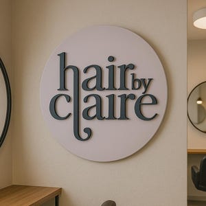 May include: A round, light pink sign with the text "hair by claire" in black lettering.