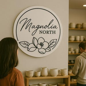 May include: A white round sign with black lettering that reads "Magnolia North". The sign features a black magnolia flower with leaves. The sign is hanging on a white wall in a shop with shelves of pottery.