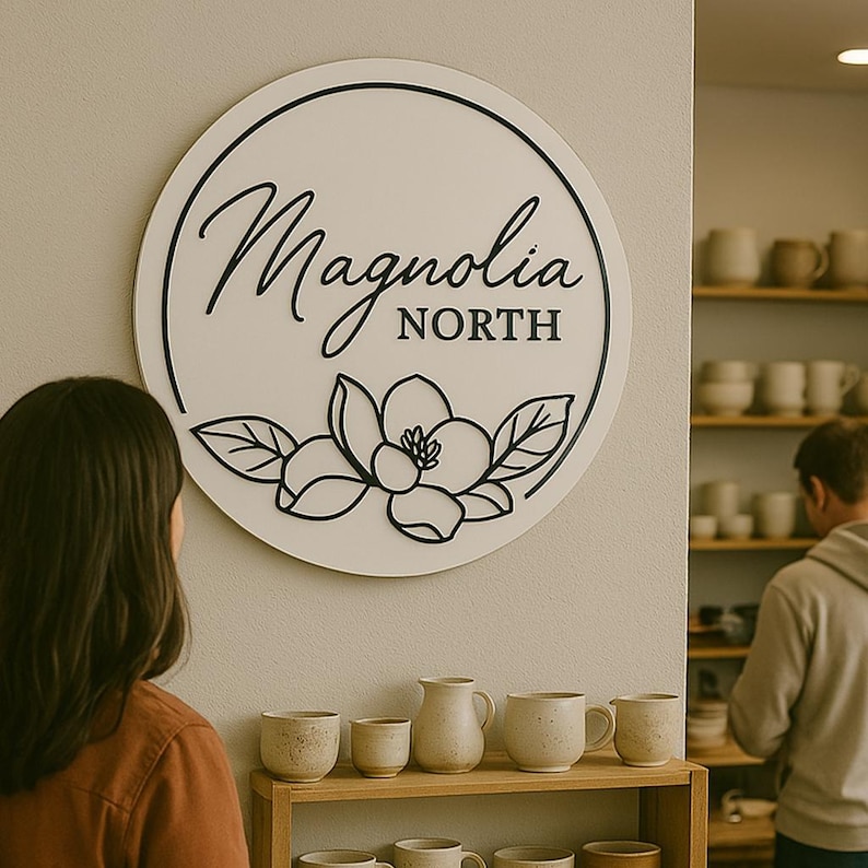 May include: A white round sign with black lettering that reads "Magnolia North". The sign features a black magnolia flower with leaves. The sign is hanging on a white wall in a shop with shelves of pottery.