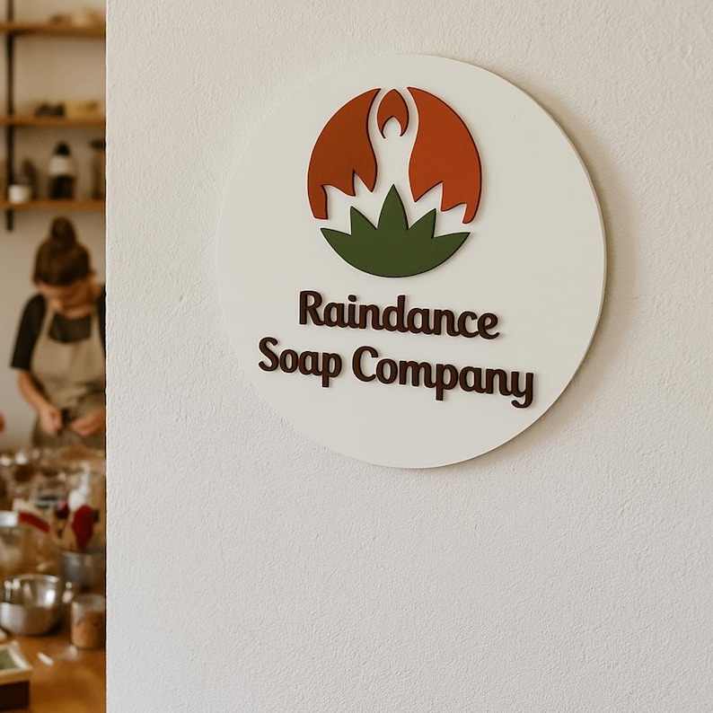 May include: A white round sign with a brown logo that says "Raindance Soap Company". The logo features a green lotus flower with a red silhouette of a person in a yoga pose.